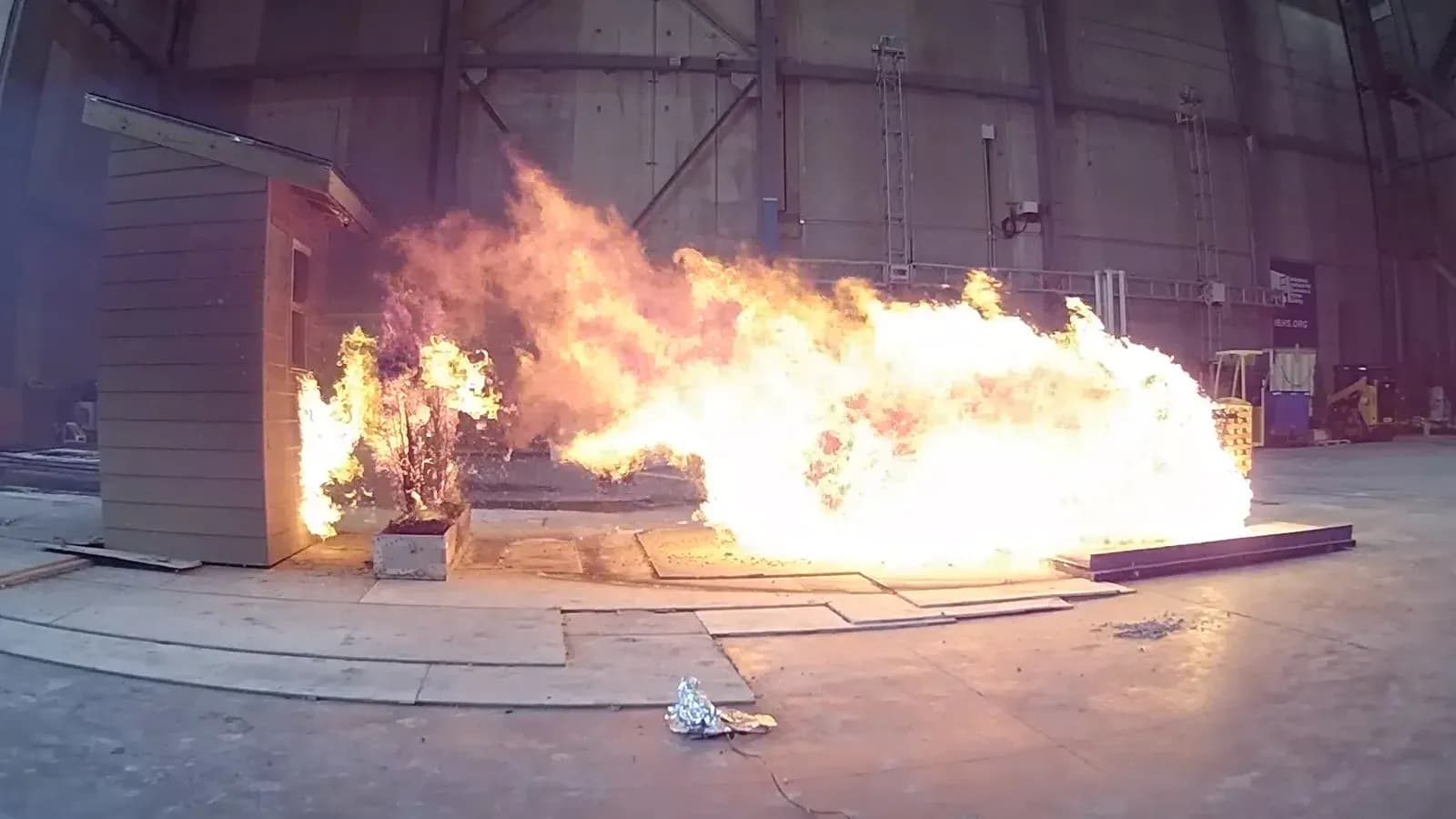 A shrub burning in a planter box against the wall of a test house during a full scale wildfire experiment inside a research laboratory.