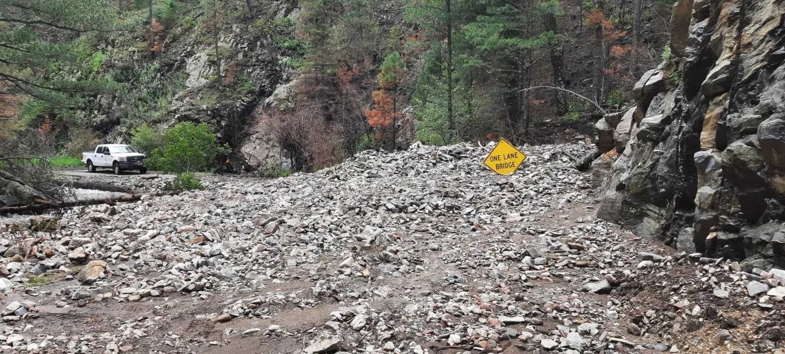 A canyon road crossing buried under a deep field of rock, gravel and mud left by a debris flow, with a road sign nearly submerged and a pickup truck parked on the clear road beyond.