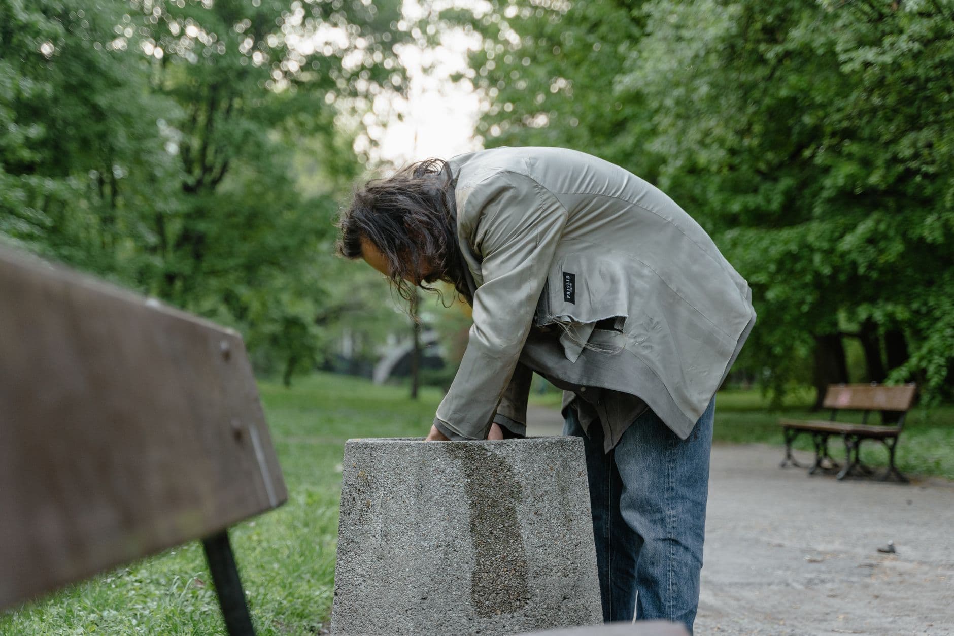 A man searches a trash bin in an urban park, highlighting urban poverty.