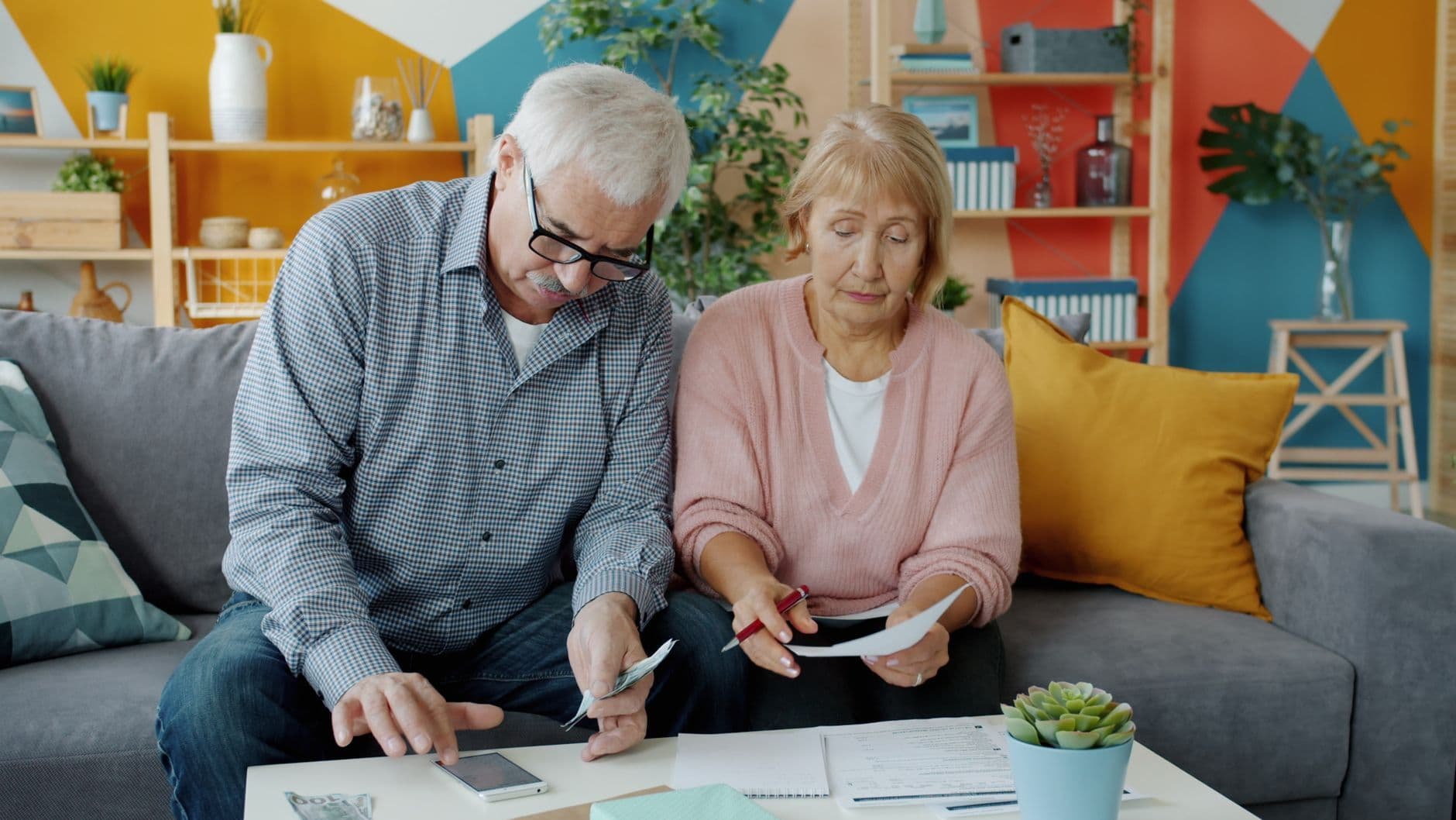 An older couple sit together on a couch at home going through household bills and cash.